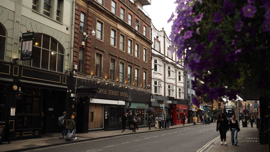 A bustling street scene in Soho featuring a row of historic brick and white buildings with shopfronts and signs, including the George Street Hotel. The street is lively with pedestrians walking along