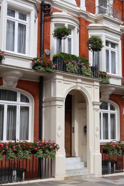 The image shows the exterior of a classic London townhouse with white decorative moldings and bay windows, set against a red brick facade. Two small potted trees with rounded green foliage are placed
