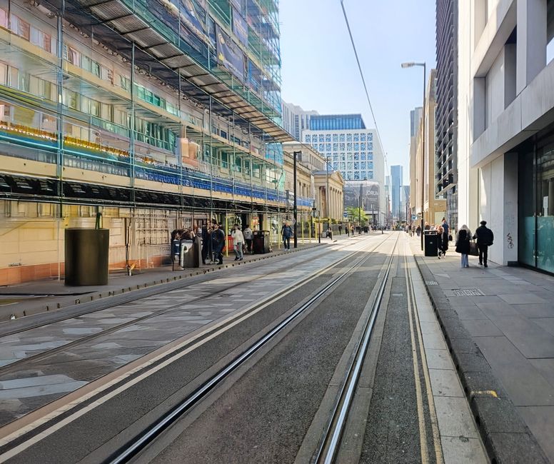 A photograph of a modern city street near Tottenham Court Road station, featuring tram tracks embedded in the asphalt running through the center. On the left, multicolored glass-fronted buildings with