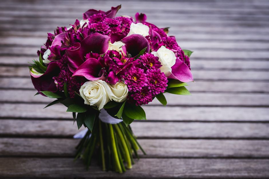 A vibrant bouquet arranged with a mix of white roses, deep purple calla lilies, and bright magenta dahlias, accented by lush green leaves. The flowers are expertly assembled in a rounded, compact styl