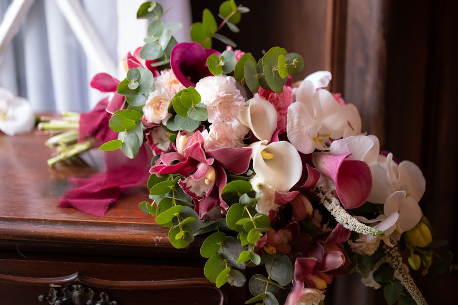 A vibrant bridal bouquet featuring a mix of white, pink, and deep magenta flowers arranged on a polished wooden surface. The bouquet includes elegant calla lilies with smooth, curved white petals, com