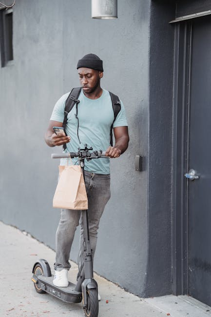 A young man with dark skin, wearing a black beanie, light blue t-shirt, and grey jeans, is standing outdoors against a grey wall. He holds a smartphone in his right hand and is balancing a brown paper