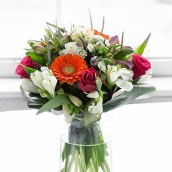 This elegant bouquet, artfully arranged and evocative of Soho's creative energy, showcases a harmony of vibrant hues and tactile contrasts. Sunset-orange gerbera daisies take center stage, their radiant petals encircling dark brown discs, while gentle peach and creamy tones soften the ensemble and evoke the subtle sunrise over Golden Square. Deep magenta roses nestle between, adding a lush sense of passion and refinement in their unfurling spirals. Delicate white alstroemeria and petite filler blossoms appear as bright, airy accents, their freshness conjuring up the lively spirit of a morning in Soho's famed Flower Market. Broad aspidistra leaves, reminiscent of the neighbourhood's understated sophistication, are expertly wrapped around the bouquet's base, their glossy surfaces forming a green cradle. Additional greenery weaves through the arrangement, enhancing its fullness and offering a medley of natural forms and textures. Set against a softly luminous backdrop, the flowers appear to glow, their colours enlivening the space, their presence radiating joyful optimism. Perfectly scaled for gifting or apartment living in Soho, this bouquet carries the artistry and attention to detail that defines the finest local florists, making it a fitting companion to special occasions, milestone moments, or heartfelt thank-yous.